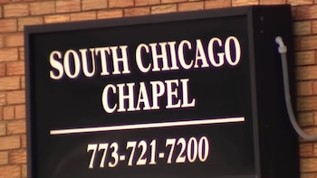 Cartel rectangular negro con texto blanco: 'SOUTH CHICAGO CHAPEL' y '773-721-7200'. El cartel está sobre una pared de ladrillos marrones