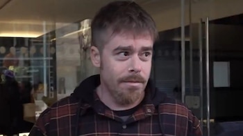 Hombre con cabello corto, barba y bigote, con perforación en la nariz, vistiendo camisa a cuadros y sudadera oscura. Fondo interior con cristales