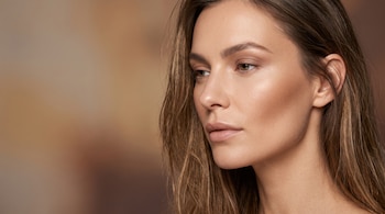 Primer plano de una mujer con maquillaje en tonos neutros, labios de color natural y sombras marrones sobre un fondo difuminado.