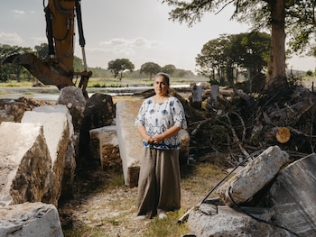 Mujer adulta, excavadora amarilla, rocas grandes, montones de escombros, ramas secas, río, árboles verdes, cielo nublado