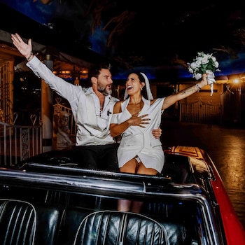 Un hombre y una mujer vestidos de novios saludan desde la parte trasera de un automóvil descapotable mientras ella sostiene un ramo de flores