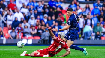 Un jugador de fútbol con uniforme rojo se desliza en el césped cerca del balón mientras otro jugador con uniforme azul lo observa.
