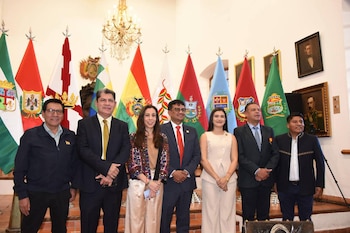 Siete personas frente a banderas de Bolivia y departamentales. Detrás, un candelabro y cuadros con retratos en la pared