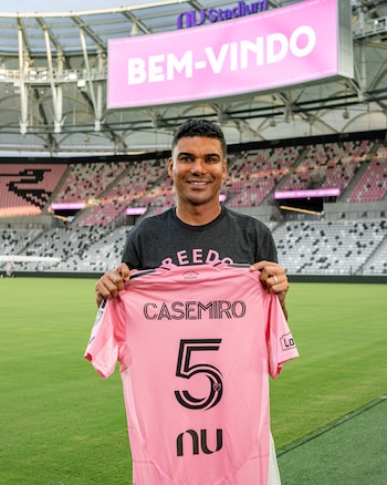 Hombre sonriente, Casemiro, sosteniendo una camiseta de fútbol rosa con el nombre Casemiro y el número cinco, en un estadio