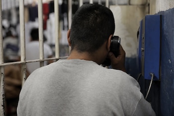 Un hombre visto de espaldas con sudadera gris habla por un teléfono de pared azul situado junto a barrotes de hierro.