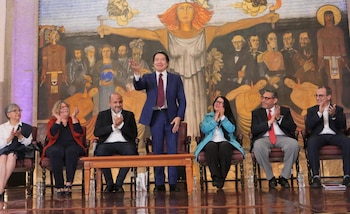 Un hombre de traje azul está de pie y saluda, rodeado por cinco personas sentadas aplaudiendo en un escenario con un gran mural histórico de fondo