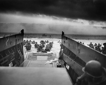 Fotografía en blanco y negro de soldados con equipo militar vadeando el agua hasta la cintura, desembarcando de una lancha de desembarco con un cielo oscuro y nublado