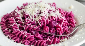 Primer plano de un plato blanco lleno de pasta en espiral de color magenta intenso, cubierta con queso parmesano rallado. Se ve un tenedor insertado en la pasta.