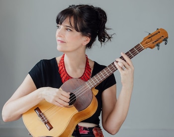 Mujer de cabello oscuro con un charango, un collar rojo y una blusa negra. La mujer mira hacia la izquierda. Fondo gris liso