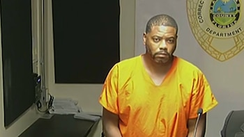 Un hombre de tez oscura con barba viste uniforme naranja. Detrás, un sello circular del 'Correctional Department', y a la izquierda, un escritorio oscuro