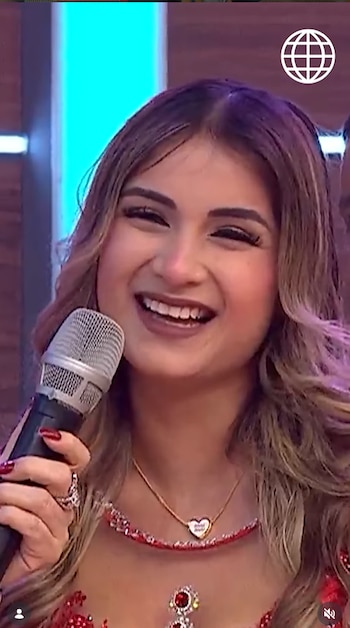 Mujer joven sonriente de cabello castaño claro sostiene un micrófono con su mano derecha. Viste un top rojo con lentejuelas y usa joyas. Hay luces de escenario