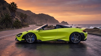 Vista lateral de un McLaren 750S Spider verde lima descapotable estacionado en una carretera mojada. Al fondo, montañas, palmeras y el mar bajo un cielo naranja al atardecer.