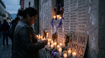 Una mujer con el cabello recogido y chaqueta oscura sostiene una vela encendida, con el rostro parcialmente oculto, frente a un muro con nombres y velas.