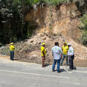 Grupo de seis personas vestidas con ropa de trabajo junto a un derrumbe de tierra y vegetación en una carretera pavimentada. Se observan herramientas