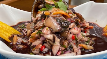 Plato blanco hondo con ceviche de conchas negras, trozos de cebolla morada, ají rojo, cilantro y líquido oscuro. Un chifle de plátano amarillo sobresale del líquido.
