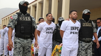 Custodios con uniforme camuflado y pasamontañas escoltan a hombres con vestimenta blanca e inscripción INPE Perú en un patio exterior.