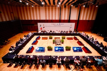 Personas sentadas alrededor de un conjunto rectangular de mesas en un auditorio iluminado con un cartel al fondo