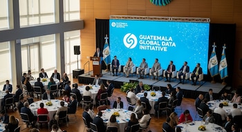 Una sala de conferencias con un podio, una gran pantalla azul que dice 'Guatemala Global Data Initiative', y varias personas sentadas en mesas redondas.
