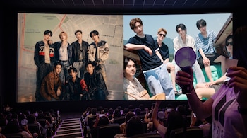Público en una sala de cine frente a una gran pantalla que exhibe dos grupos musicales mientras una persona sostiene un objeto luminoso.