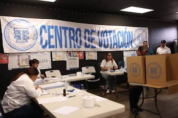Interior de un centro de votación con banner azul y blanco, varias personas en mesas y dos cabinas de votación de cartón marrón