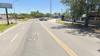 Una calle pavimentada con el texto Amadeo Sabattini pintado, un paso peatonal, una camioneta gris, árboles y carteles publicitarios