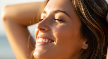 Primer plano del rostro de una mujer sonriendo con los ojos cerrados, bajo la luz del sol, con su cabello castaño y mechas claras.
