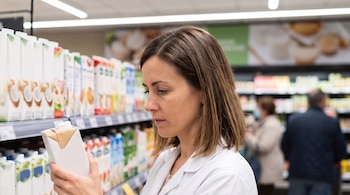 Una mujer de cabello castaño sostiene un envase de cartón blanco frente a estanterías con cartones de bebida en un supermercado.