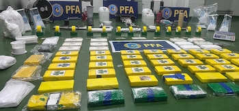 Vista aérea de una mesa verde con numerosos paquetes rectangulares de droga, algunos amarillos con tréboles, otros blancos, y algunos verdes/azules, junto a polvos blancos, balanzas y equipos