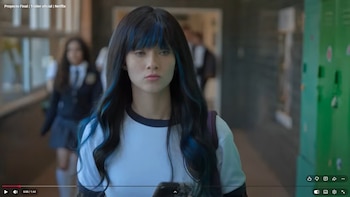 Joven con cabello negro y mechones azules, camiseta blanca, camina en pasillo con taquillas verdes. Otra estudiante en uniforme escolar al fondo