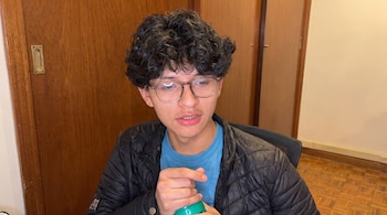 Un joven de cabello rizado con anteojos, chaqueta negra y camiseta azul sostiene un recipiente verde frente a una puerta de madera.