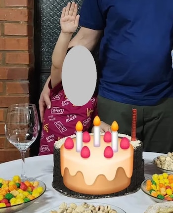 Parte de un adulto y un niño con rostro pixelado de pie junto a una mesa blanca; un pastel con cinco velas encendidas y adornos rojos, cuencos con dulces y frutos secos, y una copa vacía