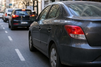 Un automóvil gris estacionado en la vía pública junto a la línea discontinua del carril mientras otros vehículos circulan al fondo.