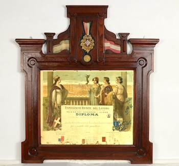 Un marco de madera tallada y oscura contiene un diploma antiguo ilustrado con figuras alegóricas, texto en italiano, una medalla y cintas de colores