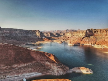 Vista aérea panorámica del Lago Powell con grandes formaciones rocosas de color rojizo a ambos lados y agua azul verdosa. Un cielo azul claro