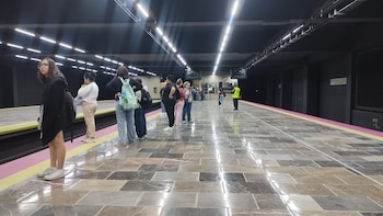 Grupo de personas de pie en un andén de metro con baldosas de mármol y franjas amarillas y rosadas, bajo un techo oscuro con luces LED, esperando un tren