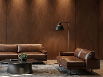 Sala moderna con tres sofás de cuero marrón, mesa de centro redonda, alfombra gris, lámpara de pie y pared de paneles de madera iluminada.