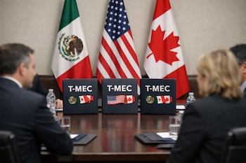 Carpetas con el logo T-MEC en una mesa de conferencias con las banderas de México, Estados Unidos y Canadá al fondo, y personas desenfocadas en los lados.