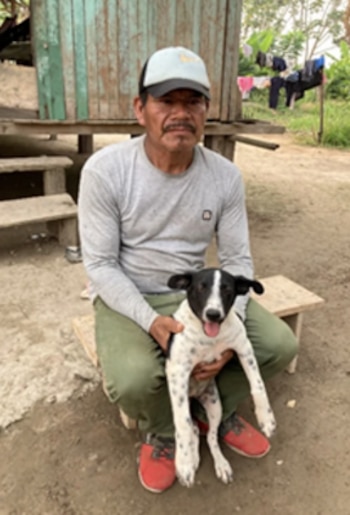 Un hombre sentado en un banco de madera sujeta a un perro blanco y negro frente a una casa de tablas