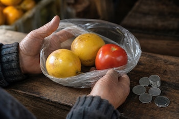 Unas manos sujetan una bolsa transparente con dos maracuyás y un tomate sobre un mostrador de madera junto a varias monedas metálicas.