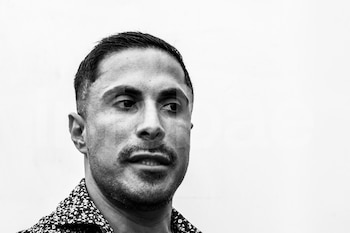 Primer plano de un hombre con cabello corto, bigote y barba corta, vestido con camisa estampada oscura sobre fondo blanco. La imagen es en blanco y negro