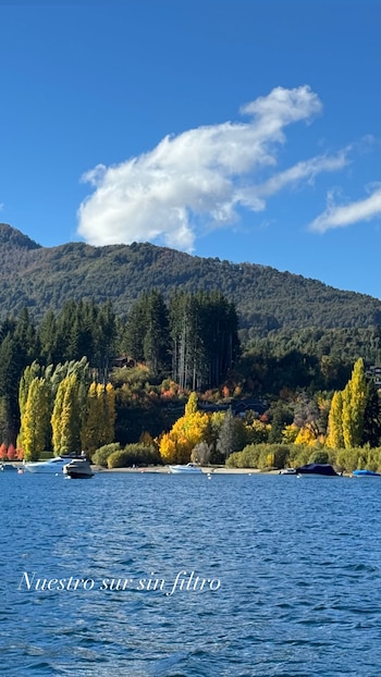 Vista panorámica de un lago azul con botes, una orilla boscosa con árboles de otoño amarillos y verdes, y una montaña bajo un cielo celeste con nubes