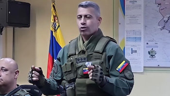 Un hombre con uniforme militar y chaleco táctico gesticula con las manos frente a una bandera de Venezuela. Hay mapas en la pared y otro hombre a la izquierda