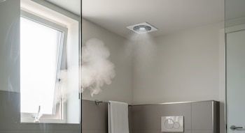 Un ventilador extractor de techo blanco expulsa vapor en un baño con paredes grises, una ventana abierta, una barra de toallas y un botón de inodoro.