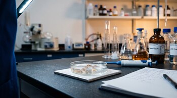 Una placa de Petri con líquido y fragmentos, una pipeta, un cuaderno con apuntes y frascos de vidrio graduados sobre una mesa de laboratorio.