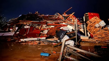 Gran pila de escombros de un edificio destruido con madera, ladrillos, metal y basura. El suelo está húmedo y oscuro bajo un cielo nocturno