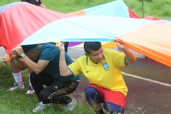 Dos hombres agachados sostienen una tela de colores azul, rojo y naranja. Uno viste camiseta amarilla de Brasil, el otro una camiseta azul. Hay césped verde
