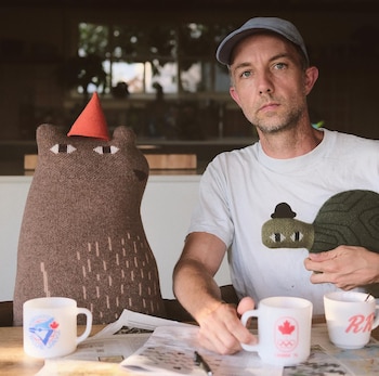 El ilustrador Jon Klassen, un hombre caucásico con gorra azul y camiseta blanca, posa con un oso de peluche marrón y una tortuga tejida en una mesa