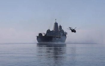 Un buque de guerra de color gris oscuro navega sobre el agua. Un helicóptero militar de color oscuro vuela al lado de la popa del buque, dejando estelas de vapor o rocío en el agua