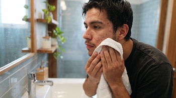 Un joven con marcas de acné en la cara se seca con una toalla blanca frente a un espejo de baño.
