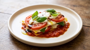 Plato de cerámica con lasaña caprese: capas de pasta, rodajas de tomate, mozzarella, albahaca, salsa de tomate, glaseado balsámico y aceite de oliva.